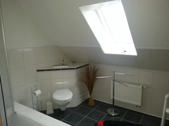 Helles Badezimmer mit Dachschräge, weißer Wand- und Bodenfliesen, Toilette, Heizkörper und großem Fenster.