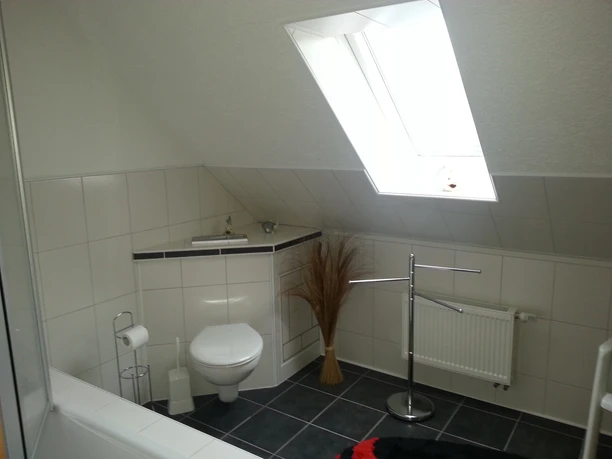 Helles Badezimmer mit Dachschräge, weißer Wand- und Bodenfliesen, Toilette, Heizkörper und großem Fenster.
