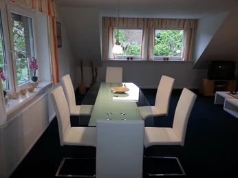 Gästewohnung Fuhrmann-Essbereich.jpg Heller Essbereich mit Glastisch und sechs modernen, weißen Stühlen vor großen Fenstern mit Blick ins Grüne.