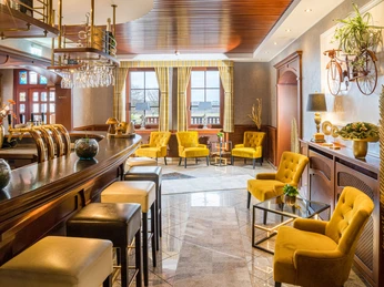 Hotel Freihof - Lobbybar.jpg Elegante Lobbybar mit gelben Sesseln, einladender Theke, Holzelementen und natürlichem Licht durch Fenster.