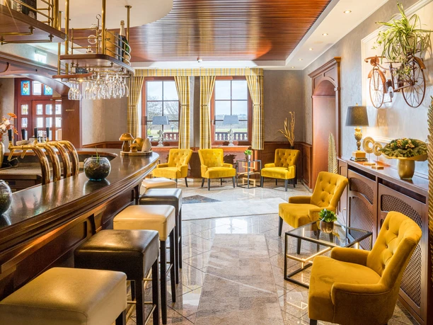 Hotel Freihof - Lobbybar.jpg Elegante Lobbybar mit gelben Sesseln, einladender Theke, Holzelementen und natürlichem Licht durch Fenster.