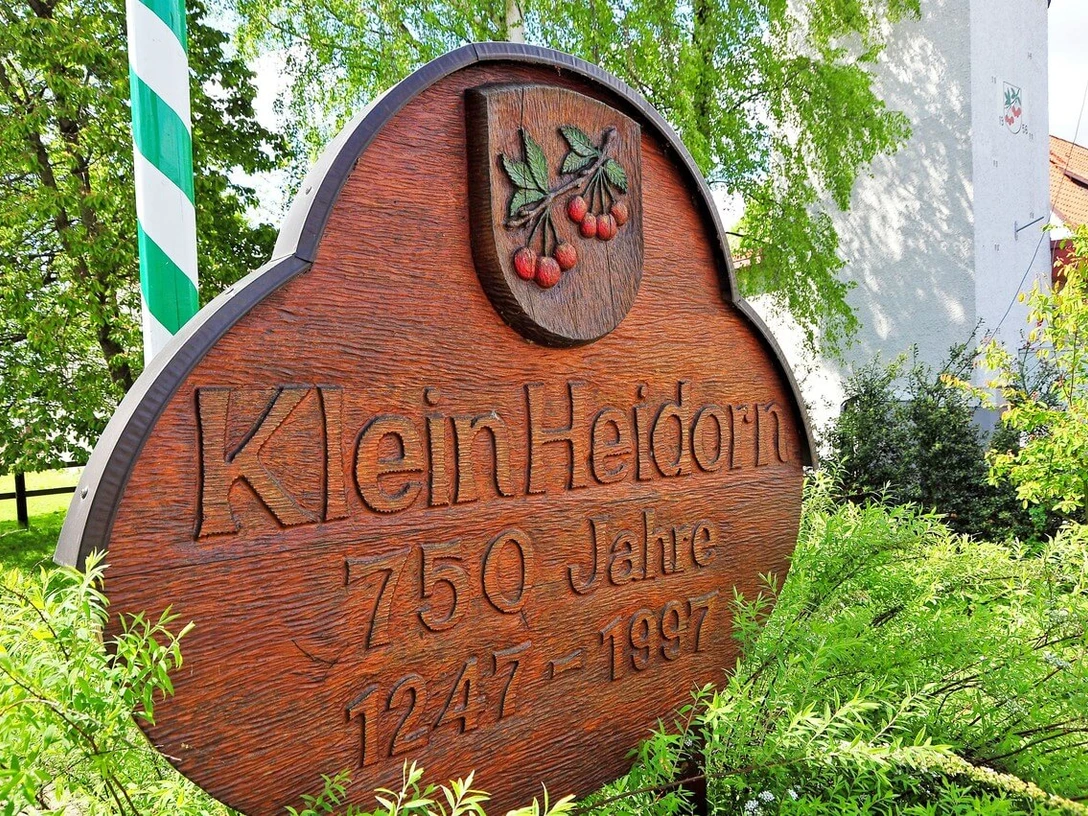 Klein Heidorn Holzschild mit der Aufschrift "Klein Heidorn 750 Jahre 1247-1997" und zwei Kirschen in grüner Umgebung.