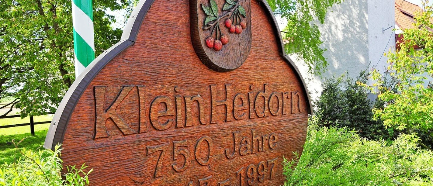 Klein Heidorn Holzschild mit der Aufschrift "Klein Heidorn 750 Jahre 1247-1997" und zwei Kirschen in grüner Umgebung.