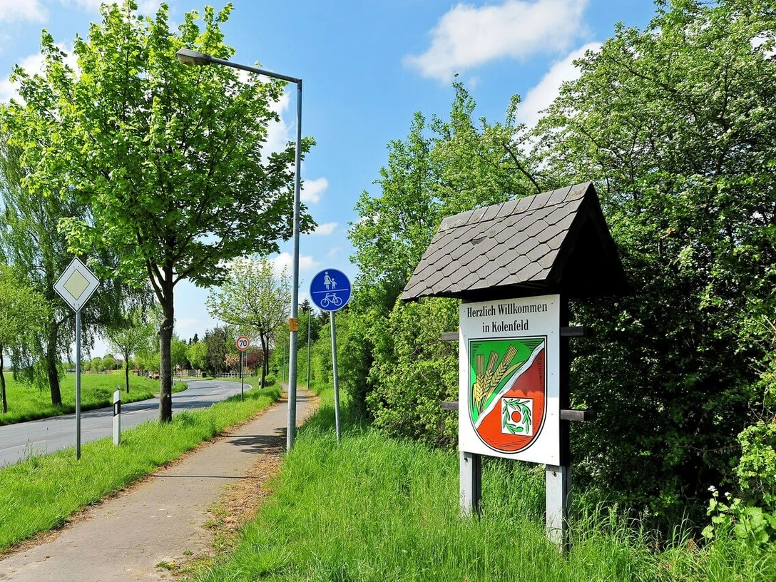Kolenfeld Ortsschild von Kolenfeld an einem sonnigen Tag, mit Radweg und grünen Bäumen im Hintergrund.