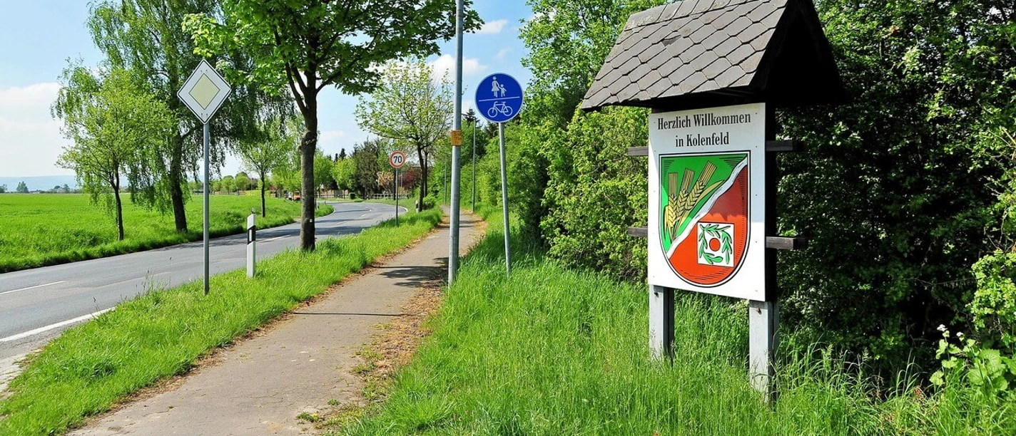 Kolenfeld Ortsschild von Kolenfeld an einem sonnigen Tag, mit Radweg und grünen Bäumen im Hintergrund.