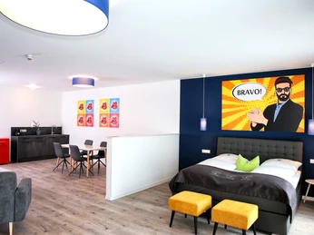 Moderne Hotel-Suite mit Bett, Sitzbereich und farbenfroher Wandkunst im Pop-Art-Stil.