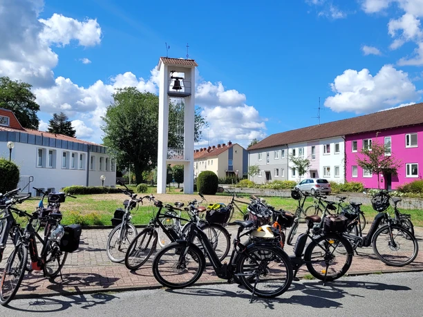 Radtouren Espelkamp Mehrere Fahrräder sind vor einem Turm mit Glocke in einer Siedlung mit bunten Gebäuden geparkt.