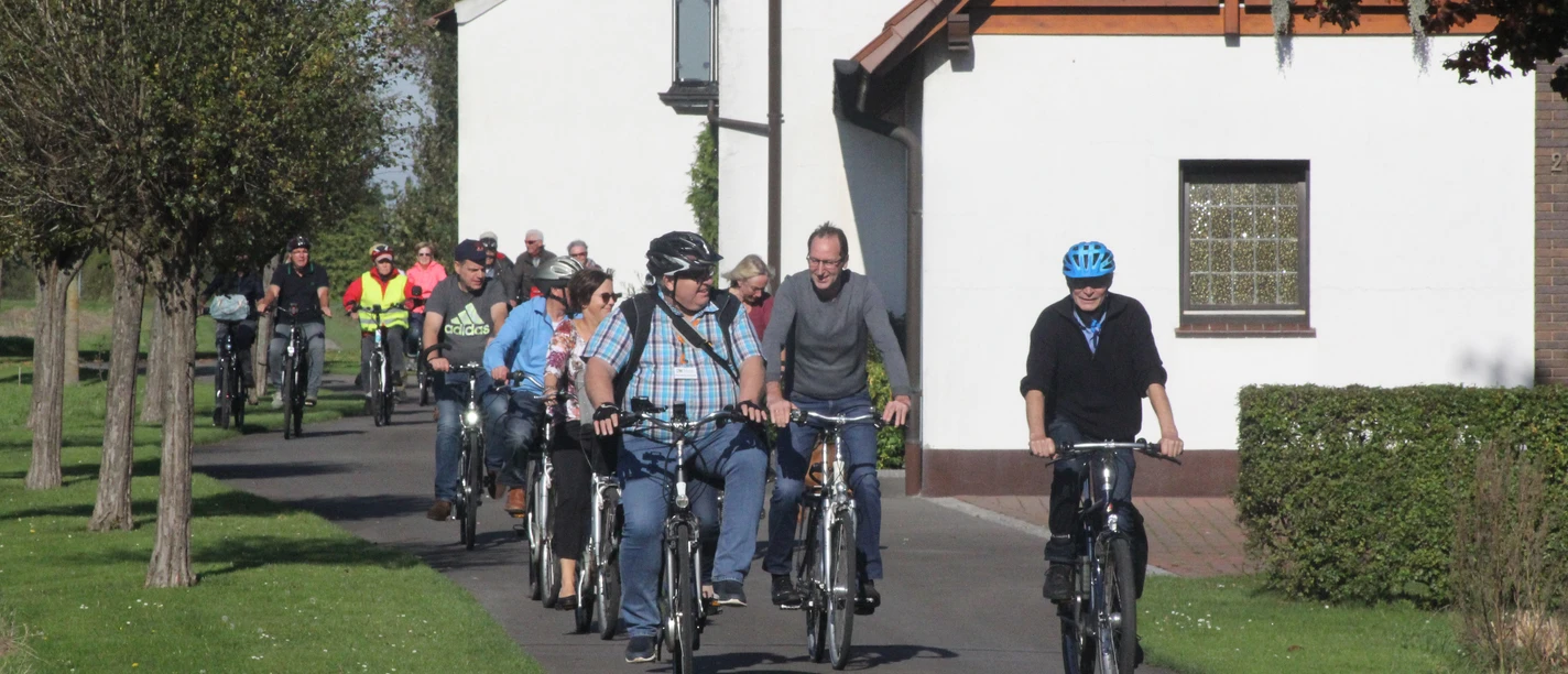 Stadtführer Espelkamp Radeln Radfahrergruppe auf asphaltiertem Weg durch ländliche Gegend, teils mit Helm und Freizeitkleidung.