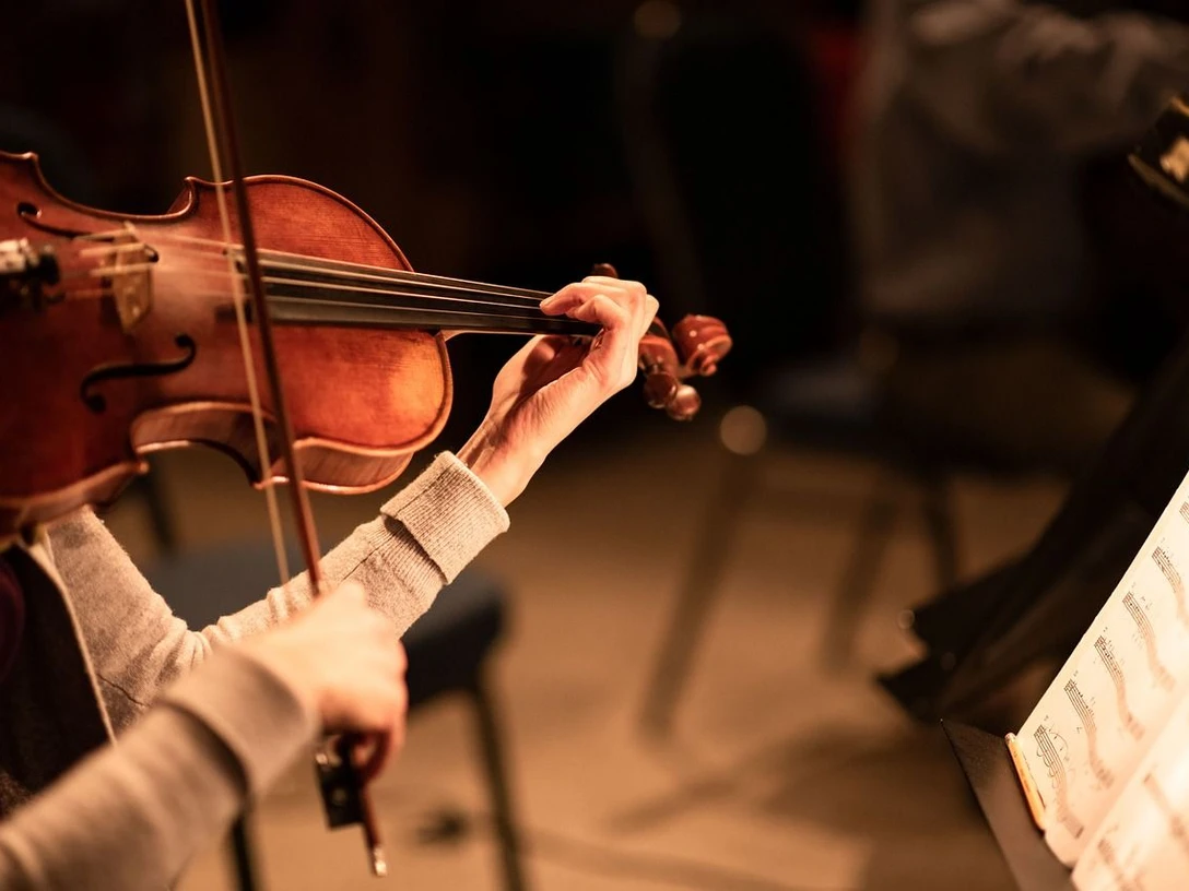Konzert_Pixabay.jpg Eine Geige wird von einer Hand festgehalten und mit einem Bogen bespielt, der von der anderen Hand festgehalten wird. Rechts steht ein aufgeschlagenes Notenbuch auf einem Notenständer.