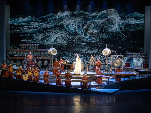 Xi'an Orchester (c) Xi'an Orchester.jpg Ein Orchester in traditionellen roten Gewändern spielt auf einer Bühne vor einem dramatischen Hintergrund.