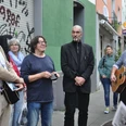 Vringsveedels tour with Wachter Loup Auf dem Bild ist eine Gruppe von Menschen unterschiedlicher Altersklassen zu sehen, die auf einer Straße in Köln steht. Zwei Männer haben Gitarren in der Hand, während ein weiterer Mann im Anzug in der Mitte steht. Einige Personen hören interessiert zu, während sie vor einer Wand mit Graffiti stehen. The picture shows a group of people of different ages standing on a street in Cologne. Two men are holding guitars, while another man in a suit stands in the middle. Some people are listening with interest as they stand in front of a wall with graffiti.