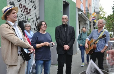 Vringsveedelsführung mit Wachter Loup Auf dem Bild ist eine Gruppe von Menschen unterschiedlicher Altersklassen zu sehen, die auf einer Straße in Köln steht. Zwei Männer haben Gitarren in der Hand, während ein weiterer Mann im Anzug in der Mitte steht. Einige Personen hören interessiert zu, während sie vor einer Wand mit Graffiti stehen.
