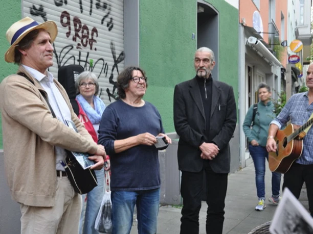 Vringsveedelsführung mit Wachter Loup Auf dem Bild ist eine Gruppe von Menschen unterschiedlicher Altersklassen zu sehen, die auf einer Straße in Köln steht. Zwei Männer haben Gitarren in der Hand, während ein weiterer Mann im Anzug in der Mitte steht. Einige Personen hören interessiert zu, während sie vor einer Wand mit Graffiti stehen.