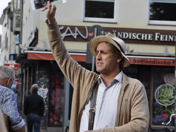 Vringsveedelsführung mit Wachter Loup Auf dem Bild ist eine Führungsperson in einem belebten Straßenzug zu sehen, die historisches Material hält.