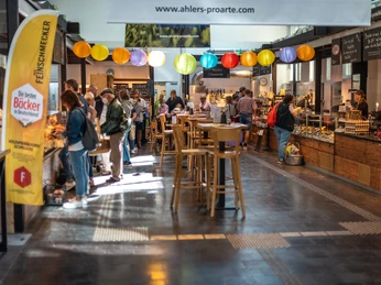 Markthalle_Herford_6.8.22_042.jpg Menschen schlendern durch eine belebte Markthalle mit bunten Lampions und Ständen voller Köstlichkeiten.
