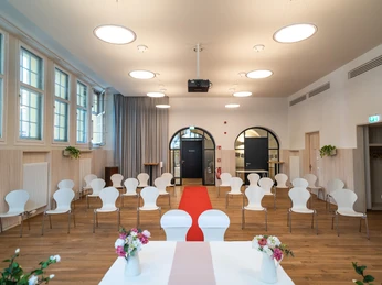 Ein festlich hergerichteter Raum mit weißen Stühlen, Blumenarrangements und rotem Teppich.