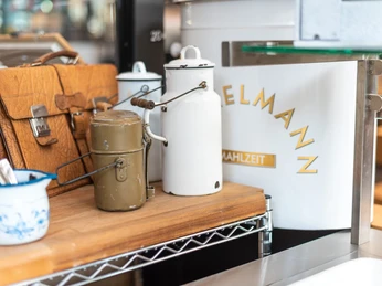 Markthalle_Herford_2022_287.jpg Traditionelle Milchkanne und Brotdose aus Metall auf einem Holztisch in einer Markthalle platziert.