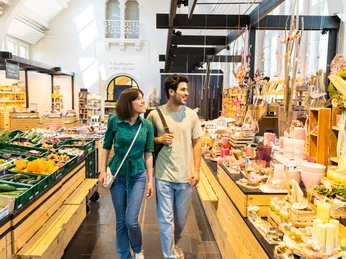 herford-markthalle-teutoburger-wald-tourismus-h-tornow-009-jpg.jpg Eine Marktbesucherin und ein Besucher schlendern lächelnd durch die bunte Herforder Markthalle.