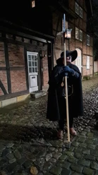 Ein Nachtwächter in historischem Gewand mit Laterne steht auf einem Kopfsteinpflaster vor Fachwerkhäusern.