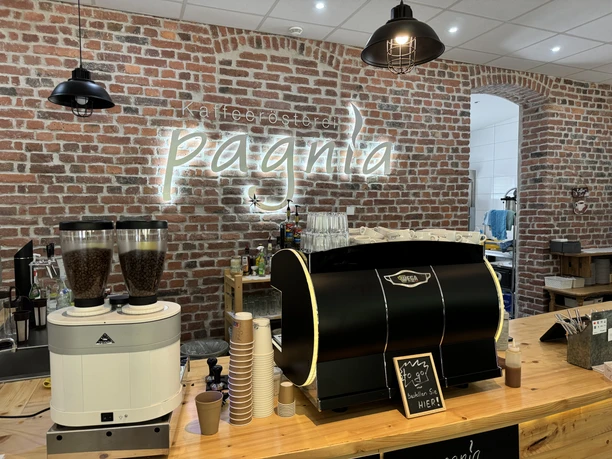 Kaffeerösterei Pagnia - Ansicht Theke In einem Café mit Ziegelmauer optik stehen Kaffeemaschinen und Pappbecher auf einer Holztheke.