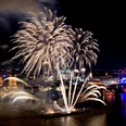 Fireworks burst above a riverside cityscape at night, reflected in the water beneath.