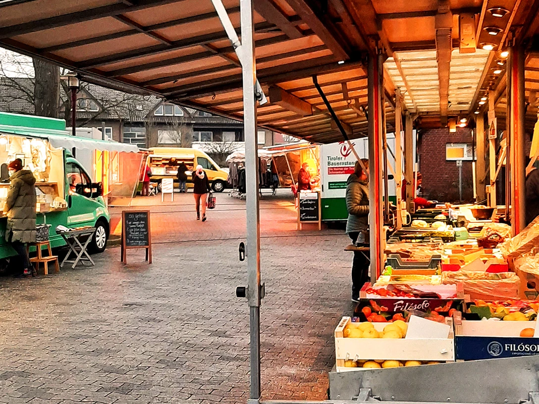 Obst- und Gemüsestände auf einem typischen Wochenmarkt bei Tageslicht mit Besuchern.