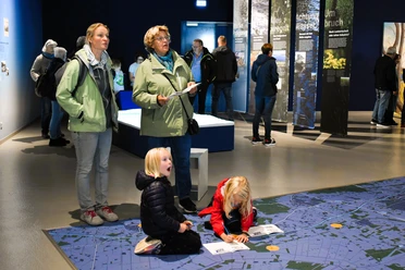 Weihnachtlicher Familiennachmittag_nbz-Alfsee.jpg Drie volwassenen kijken naar spelende kinderen op een grote blauwe plattegrond in een tentoonstellingsruimte.
