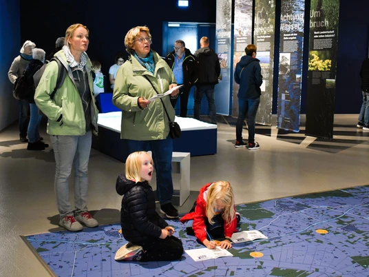 Weihnachtlicher Familiennachmittag_nbz-Alfsee.jpg Drei Erwachsene beobachten Kinder, die auf einem großen, blauen Bodenplan in einem Ausstellungsraum spielen.Three adults watch children playing on a large blue floor plan in an exhibition room.Tre voksne ser på børn, der leger på en stor blå gulvplan i et udstillingsrum.Drie volwassenen kijken naar spelende kinderen op een grote blauwe plattegrond in een tentoonstellingsruimte.