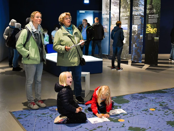 Weihnachtlicher Familiennachmittag_nbz-Alfsee.jpg Drei Erwachsene beobachten Kinder, die auf einem großen, blauen Bodenplan in einem Ausstellungsraum spielen.Three adults watch children playing on a large blue floor plan in an exhibition room.Tre voksne ser på børn, der leger på en stor blå gulvplan i et udstillingsrum.Drie volwassenen kijken naar spelende kinderen op een grote blauwe plattegrond in een tentoonstellingsruimte.