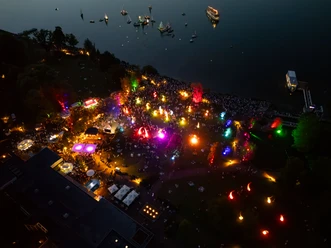 lichternacht-von-oben.jpg Aus der Vogelperspektive zeigt das Bild die lebhafte Veranstaltung Lichternacht bei Nacht, erhellt durch bunte Lichter, mit einem angrenzenden dunklen Wasserbereich und vereinzelten kleinen Booten.