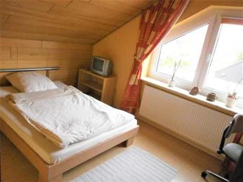 Ein kleines Schlafzimmer mit Einzelbett, Dachschräge, Fenster, rotem Vorhang und einem TV-Gerät.