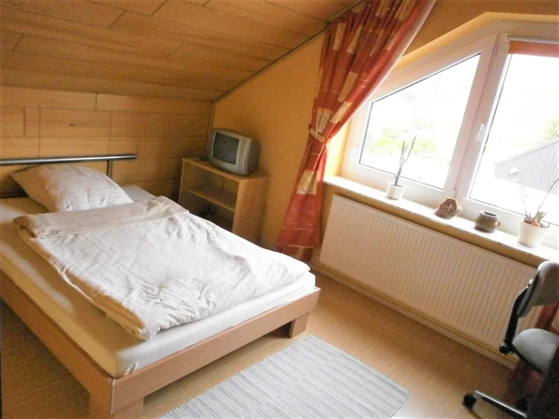 FeWo-Koetter-kleines Schlafzimmer.jpg Ein kleines Schlafzimmer mit Einzelbett, Dachschräge, Fenster, rotem Vorhang und einem TV-Gerät.
