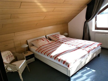 FeWo-Koetter-Schlafraum.jpg Zimmer mit Doppelbett, Holzverkleidung an der Decke, Fenster mit Vorhängen, Korbsessel, Lampe.