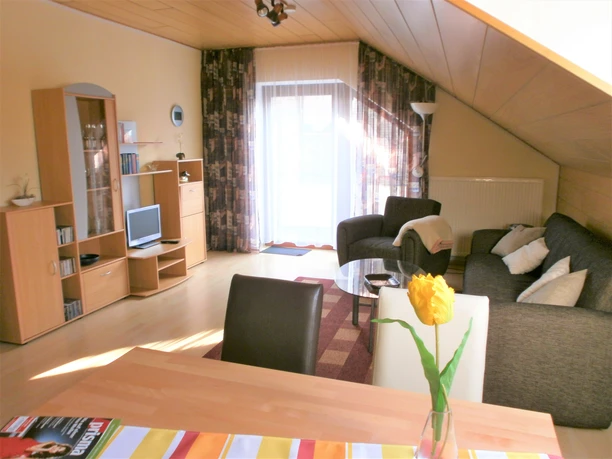 Wohnraum mit Sofa, Sessel, Flachbild-TV und Esstisch, helle Einrichtung und Zugang zu einem Balkon.