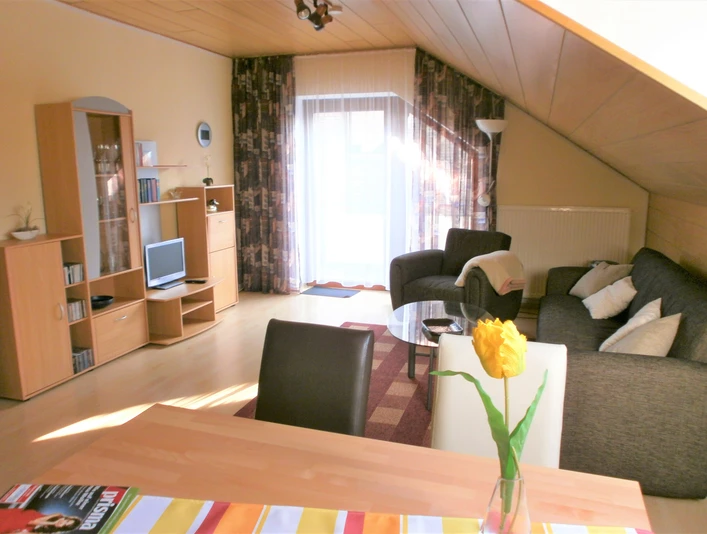 Wohnraum mit Sofa, Sessel, Flachbild-TV und Esstisch, helle Einrichtung und Zugang zu einem Balkon.