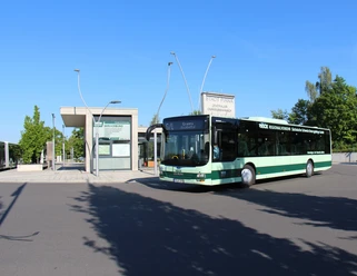 Een groene bus staat voor een modern bushaltegebouw bij zonnig weer; op de achtergrond zijn bomen en een blauwe lucht te zien.