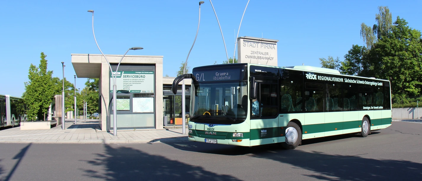 Een groene bus staat voor een modern bushaltegebouw bij zonnig weer; op de achtergrond zijn bomen en een blauwe lucht te zien.