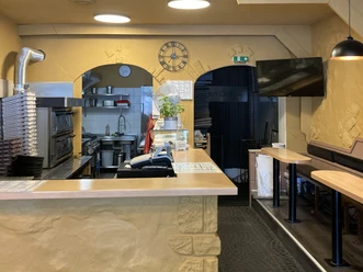 Innenansicht der Pizzeria mit Theke, Pizzaöfen, Stapel von Pizzakartons und Tischen auf der rechten Seite.