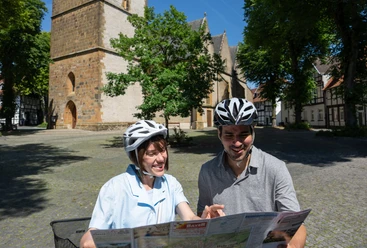 halle-kirchplatz-teutoburger-wald-tourismus-patrick-gawandtka-081-jpg_quer.jpg Ein lächelndes Paar mit Fahrradhelmen betrachtet eine Landkarte vor einer historischen Kirche.
