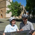 halle-kirchplatz-teutoburger-wald-tourismus-patrick-gawandtka-081-jpg_quer.jpg Ein lächelndes Paar mit Fahrradhelmen betrachtet eine Landkarte vor einer historischen Kirche.