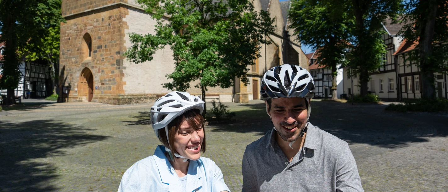 halle-kirchplatz-teutoburger-wald-tourismus-patrick-gawandtka-081-jpg_quer.jpg Ein lächelndes Paar mit Fahrradhelmen betrachtet eine Landkarte vor einer historischen Kirche.