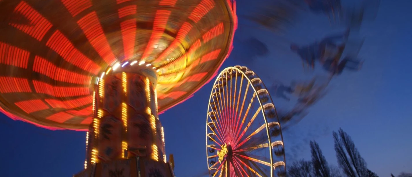 Ein beleuchtetes Karussell dreht sich neben einem erleuchteten Riesenrad bei Dämmerung auf dem Aachener Bend.