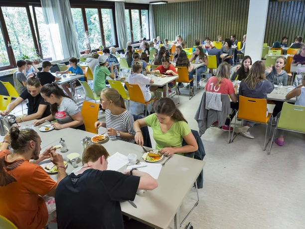 Jugendherberge Lindlar Eine große Gruppe junger Menschen sitzt in einer gut beleuchteten Cafeteria an mehreren Tischen.