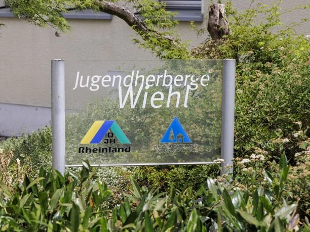 Jugendherberge Wiehl Schild der Jugendherberge Wiehl inmitten üppiger grüner Sträucher und Pflanzen vor einem Gebäude.