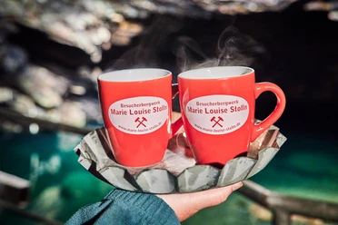 Twee rode kopjes met het logo van "Besucherbergwerk Marie Louise Stolln", poserend voor de achtergrond van een grot.