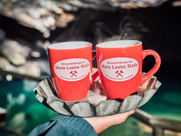 Glühwein am See Zwei rote Tassen mit "Besucherbergwerk Marie Louise Stolln"-Logo, vor einer Höhlenkulisse posierend.