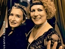 Zwei Frauen in eleganter Abendgarderobe lächeln, geschmückt mit Federn und Perlen. Sie posieren zusammen.