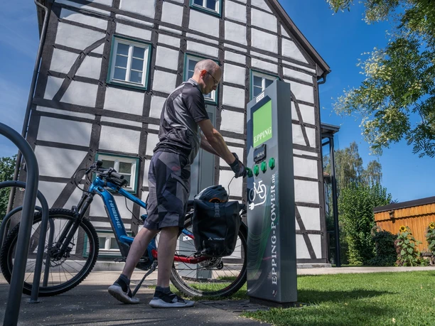 Person lädt E-Bike an moderner Station vor Fachwerkgebäude unter klarem Himmel in Hövelhof auf.