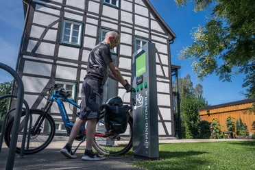 E-Bike-Ladestation an der Tourist-Info Hövelhof Person lädt E-Bike an moderner Station vor Fachwerkgebäude unter klarem Himmel in Hövelhof auf.