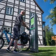 E-Bike-Ladestation an der Tourist-Info Hövelhof Person lädt E-Bike an moderner Station vor Fachwerkgebäude unter klarem Himmel in Hövelhof auf.